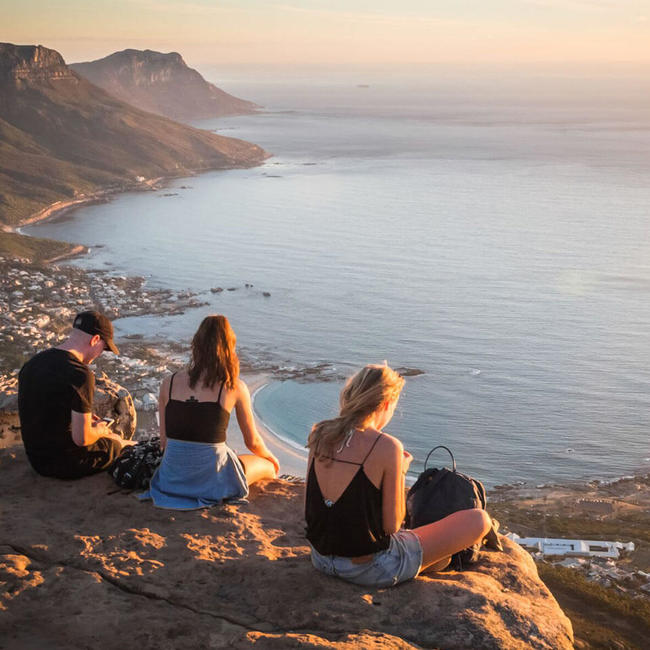 three young adults sitting on cliff watching sunset in cape town